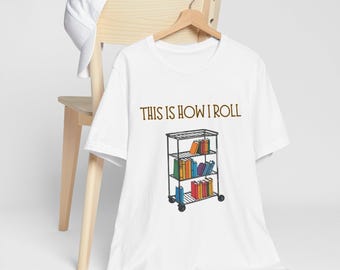 Op de afbeelding: Wit T-shirt met een bruine afbeelding van een boekenkast met kleurrijke boeken en de tekst "This is how I roll".