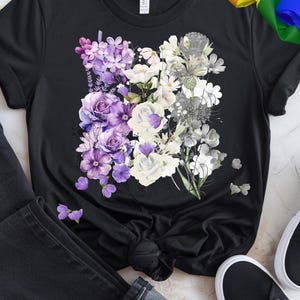 May include: Black t-shirt featuring a floral design with purple, white, and gray flowers. The design includes roses, hydrangeas, and other blooms. The shirt is paired with black jeans and black sneakers.