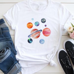 Puede incluir: Camiseta blanca con un diseño de acuarela de planetas en tonos naranja, rojo, azul y verde.