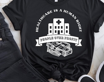 May include: Black t-shirt with white text that reads "Healthcare is a human right. People over profit." The text is in a circle around a white silhouette of a hospital building. Below the building is a stack of money with a banner that reads "People over profit."