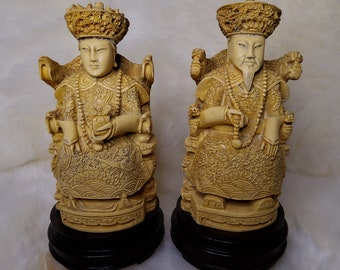 Op de afbeelding: Twee ivoorwitte beeldjes van Chinese royalty, elk zittend op een troon met ingewikkelde houtsnijwerk. De figuren dragen uitgebreide gewaden en hoofdtooien, en ze houden voorwerpen in hun handen.