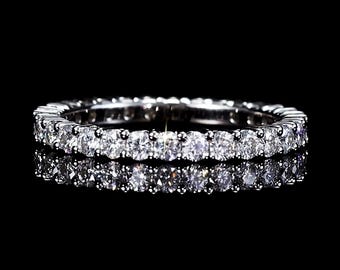 May include: A silver eternity ring with a continuous row of round, clear gemstones. The ring is set against a black background, with a reflection visible below the ring.