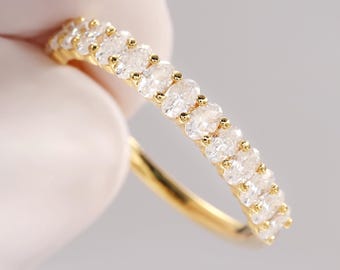 May include: A gold ring featuring a row of oval-cut, clear gemstones. The ring's band is a polished gold color, and the gemstones are set closely together. The ring is held against a white background.