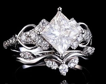May include: A silver-toned ring set featuring a large princess-cut center stone flanked by smaller pavé-set diamonds. The band displays intricate, swirling metalwork and leaf-like accents, creating a layered appearance with a reflective finish.