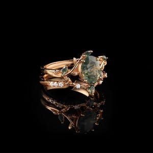 May include: A rose gold ring set featuring an oval moss agate gemstone, surrounded by smaller green gemstones and delicate leaf and vine detailing. The set includes a band with small, clear gemstones. The rings are displayed on a reflective black surface.