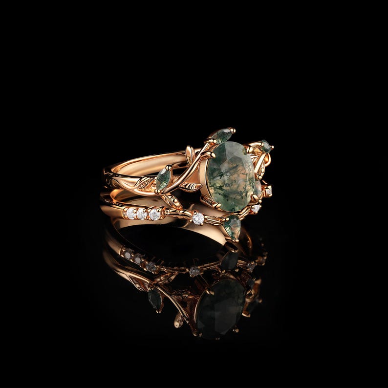 May include: A rose gold ring set featuring an oval moss agate gemstone, surrounded by smaller green gemstones and delicate leaf and vine detailing. The set includes a band with small, clear gemstones. The rings are displayed on a reflective black surface.