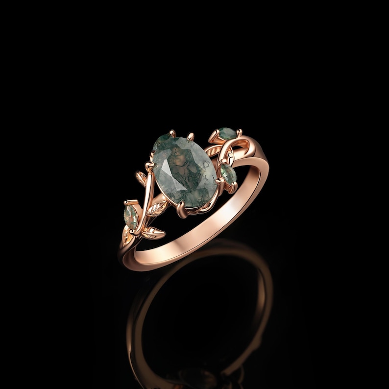 May include: Rose gold ring featuring an oval moss agate gemstone. The ring is adorned with delicate leaf and small gemstone accents. The design is set against a black background, highlighting the ring's intricate details and natural stone.