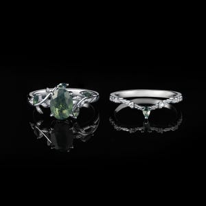 May include: Two silver rings. One ring features a large, oval-cut green gemstone with a leafy vine design. The other ring is a silver band with small clear gemstones and a small green gemstone.