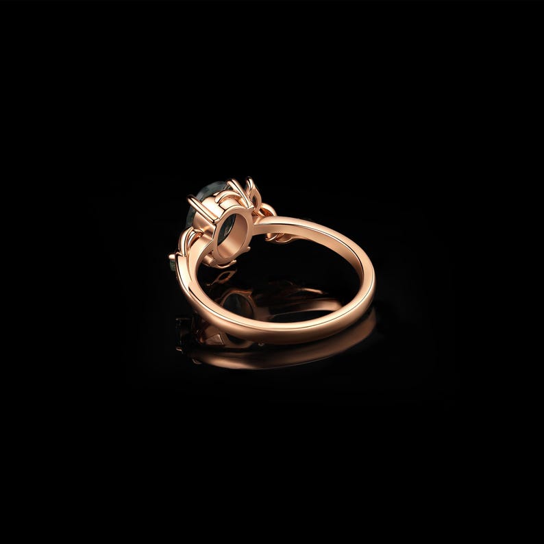 May include: Rose gold ring with a dark teal oval gemstone. The ring features a detailed floral design around the gemstone and on the band. The ring is set against a black background, highlighting its elegant design.