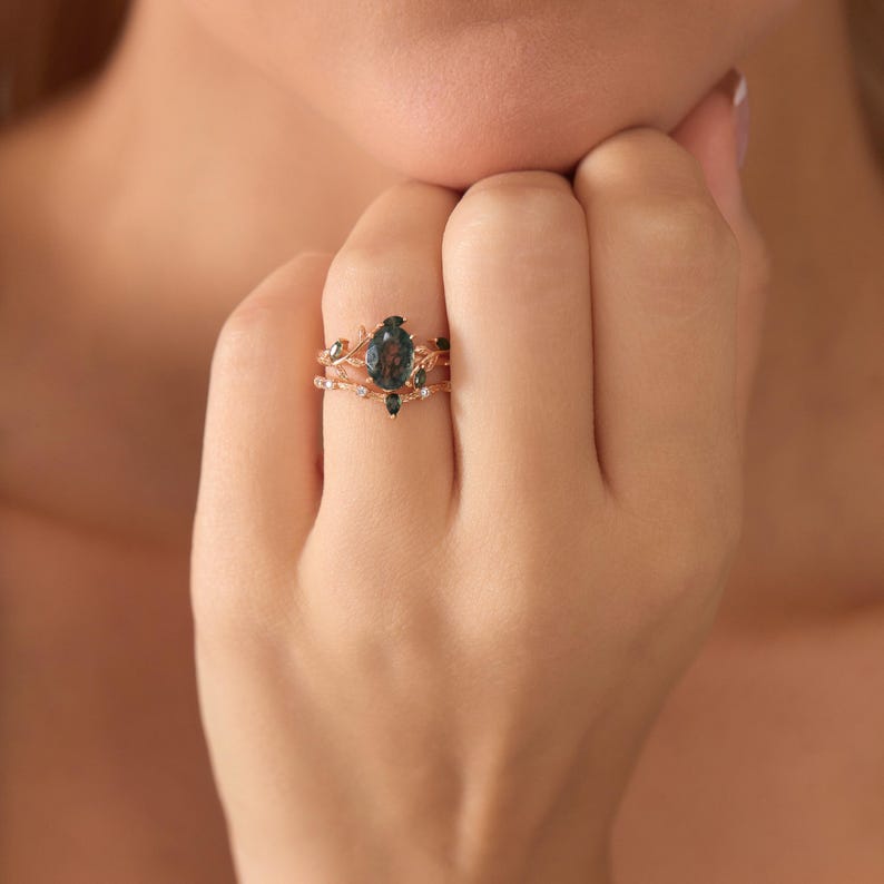 May include: A rose gold ring set featuring a large, dark green teardrop-shaped gemstone as the centerpiece. The ring is accented with smaller green and white gemstones and delicate leaf-like designs.