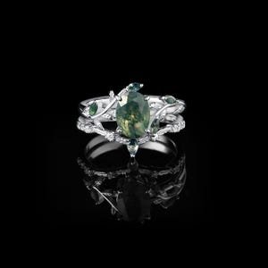 May include: A silver ring set with a pear-shaped green moss agate gemstone and smaller green accent stones. The ring features a delicate vine and leaf design, with a matching band. The set is displayed on a reflective black surface.