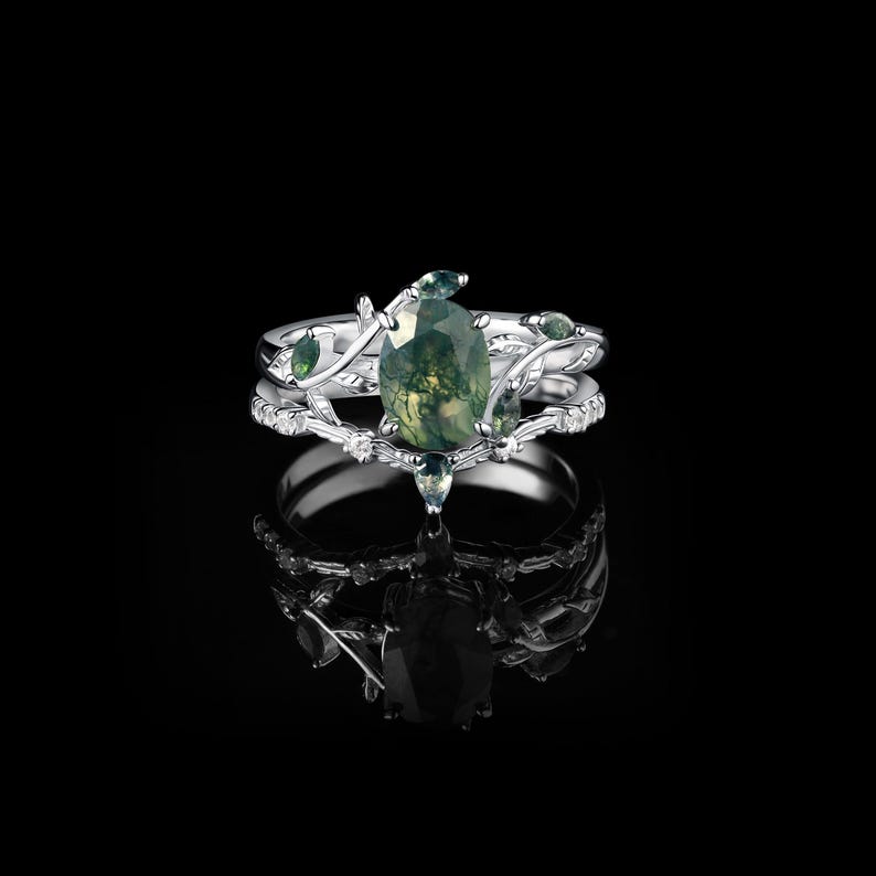 May include: A silver ring set with a pear-shaped green moss agate gemstone and smaller green accent stones. The ring features a delicate vine and leaf design, with a matching band. The set is displayed on a reflective black surface.