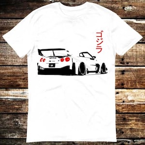 Puede incluir: Camiseta blanca con un gráfico en blanco y negro de un coche deportivo y luces traseras rojas. La palabra japonesa "Godzilla" está impresa en rojo en el lado derecho. La camiseta está hecha de un material suave y cómodo.