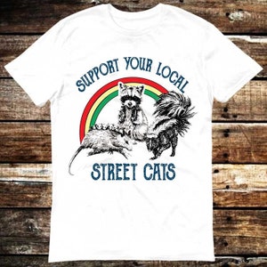 May include: White t-shirt featuring a rainbow graphic and the text "Support Your Local Street Cats". The graphic depicts a raccoon, a skunk, and a possum.