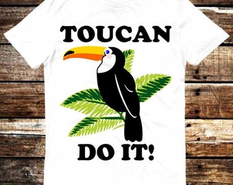 Puede incluir: Camiseta blanca con una ilustración de tucán negro y hojas verdes. El tucán tiene un pico amarillo y ojos azules. El texto "TOUCAN DO IT!" está impreso en negro.