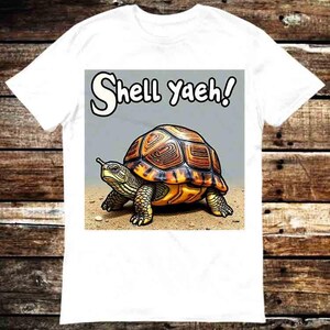 以下が含まれることがあります： 白いTシャツに、茶色と黄色の甲羅を持つカメの漫画のイラストが描かれています。カメは砂の上に立っています。「Shell Yaeh!」というテキストがカメの上にあります。Tシャツは白い生地で作られています。
