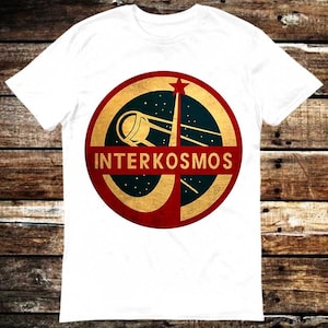 May include: White t-shirt with a circular design that reads "INTERKOSMOS". The design incorporates a gold ring, a red star, and a stylized illustration of a satellite or spacecraft against a dark blue backdrop.