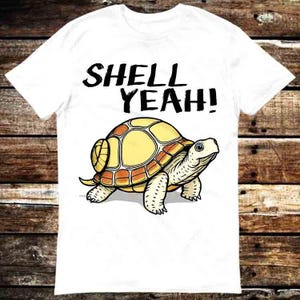 May include: White t-shirt with a cartoon turtle illustration. The turtle has a yellow shell with orange accents and is walking. The text "SHELL YEAH!" is above the turtle in black lettering. The shirt is on a wooden background.