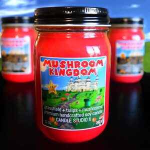 May include: Three red glass candle jars with black lids. The center jar has a label that reads "Mushroom Kingdom" with a cartoon image of a castle and a star. The label also says "grassfield + tulips + mushrooms premium handcrafted soy candle 12 oz Candle Studio X 340g".