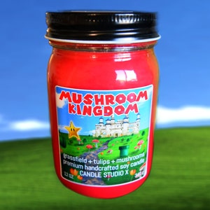 May include: A red glass jar candle with a black lid and a label that says "Mushroom Kingdom" with a cartoon image of a castle, a star, and mushrooms. The label also says "grassfield + tulips + mushrooms premium handcrafted soy candle 12 oz CANDLE STUDIO X".