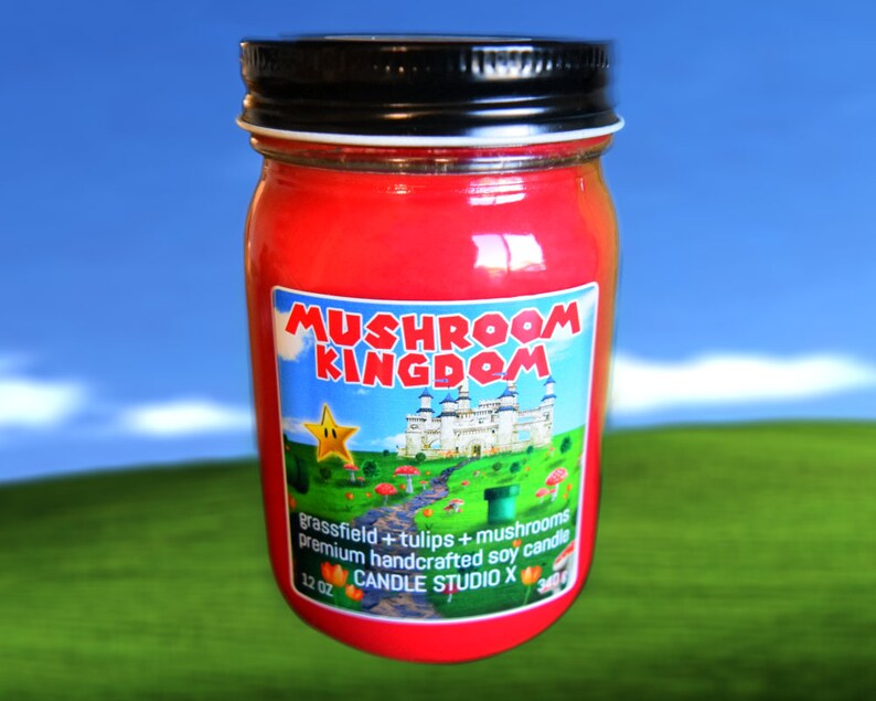 May include: A red glass jar candle with a black lid and a label that says "Mushroom Kingdom" with a cartoon image of a castle, a star, and mushrooms. The label also says "grassfield + tulips + mushrooms premium handcrafted soy candle 12 oz CANDLE STUDIO X".