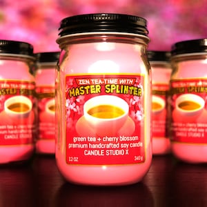 May include: Three pink glass jars with black lids and labels. The center jar has a label that reads "Zen Tea-Time with Master Splinter" and "green tea + cherry blossom premium handcrafted soy candle Candle Studio X 12 oz 340 g".