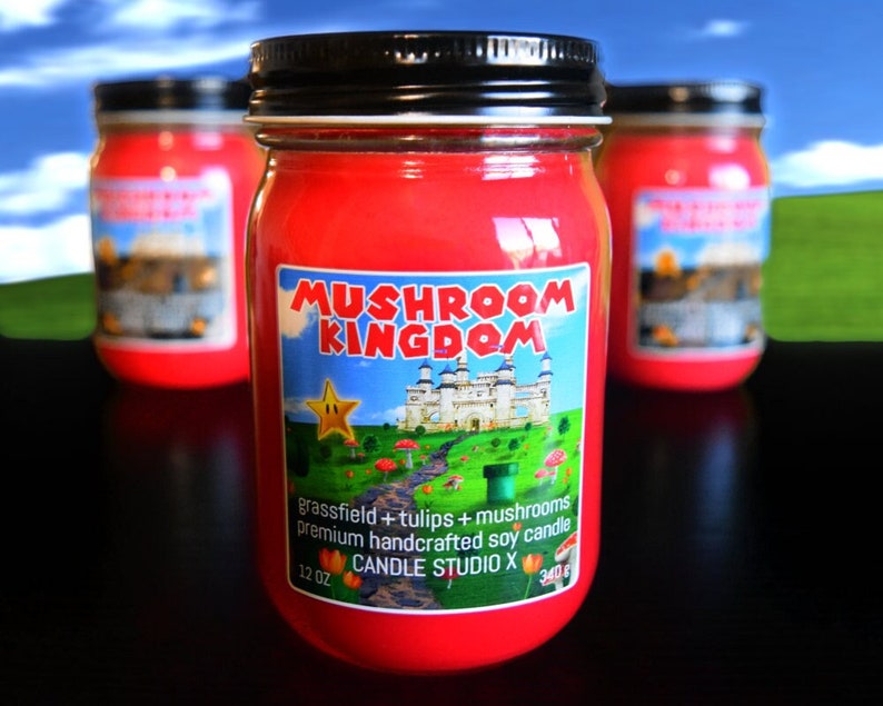 May include: Three red glass candle jars with black lids. The center jar has a label that reads "Mushroom Kingdom" with a cartoon image of a castle and a star. The label also says "grassfield + tulips + mushrooms premium handcrafted soy candle 12 oz Candle Studio X 340g".