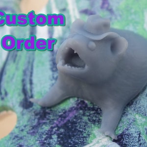 Op de afbeelding: Een grijs 3D-geprint beeldje van een klein dier dat een cowboyhoed draagt. Het beeldje heeft zijn mond open en lijkt te schreeuwen. Het beeldje staat op een kleurrijk oppervlak met de woorden "Custom Order" in paarse tekst.