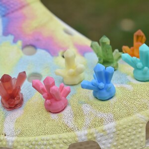 Op de afbeelding: Een close-up van zeven kleurrijke kristallen schoenbedels. De bedels zijn in de vorm van kristallen en zijn gemaakt van plastic. De bedels zijn in de kleuren rood, roze, geel, blauw, groen en oranje.