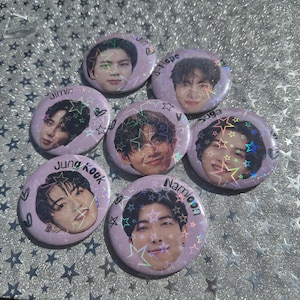 May include: Seven circular pin-back buttons featuring portraits of individuals with holographic star patterns overlaying the images. Each button displays a name printed in black text, including Jin, J-Hope, Jimin, V, Suga, Jung Kook, and Namjoon.