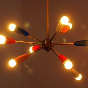 May include: A gold-toned, mid-century modern chandelier with six arms, each with a different coloured tip. The tips are red, blue, white, and orange. The chandelier has six light bulbs that are turned on and glowing.