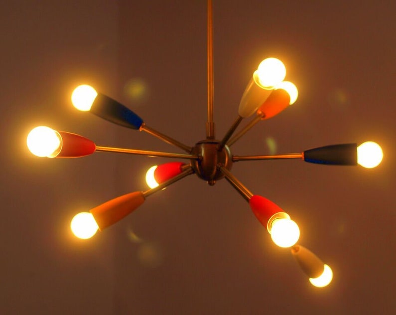 May include: A gold-toned, mid-century modern chandelier with six arms, each with a different coloured tip. The tips are red, blue, white, and orange. The chandelier has six light bulbs that are turned on and glowing.