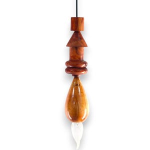 May include: A handcrafted wooden pendant light fixture. The fixture features a series of geometric shapes, including a square, a triangle, and rounded elements, all made from polished wood. A teardrop-shaped element with a white, flame-like glass piece at the bottom completes the design.