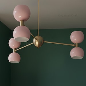 Könnte beinhalten: Eine moderne Deckenleuchte mit Messing-Finish und sechs rosa kugelförmigen Leuchten. Die Leuchten sind paarweise angeordnet und an einer zentralen Halterung aufgehängt. Der Text "Kingsman Craft" ist im Hintergrund sichtbar.