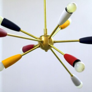 May include: A gold-toned, multi-armed chandelier with coloured accents. The arms extend outwards from a central sphere, each ending in a light bulb with a coloured shade. The shades are red, yellow, blue, white, and grey.