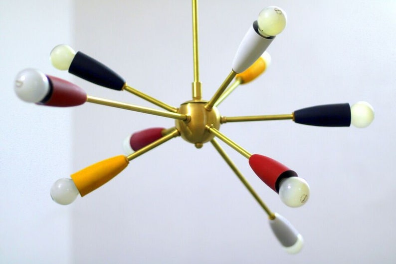 May include: A gold-toned, multi-armed chandelier with coloured accents. The arms extend outwards from a central sphere, each ending in a light bulb with a coloured shade. The shades are red, yellow, blue, white, and grey.
