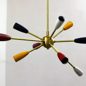 May include: A brass and multi-coloured mid-century modern chandelier with eight light bulbs. The chandelier has a starburst design with arms extending out from a central sphere. The light bulbs are covered in shades of red, yellow, white, and black.