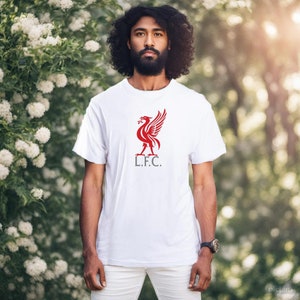 May include: White t-shirt featuring a red bird emblem and the letters "L.F.C." The short-sleeved shirt is worn by a person. The person is wearing white trousers and a watch. The background is a green bush.