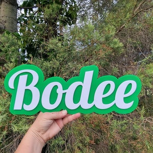 Op de afbeelding: Een groen en wit gepersonaliseerd naambord met de naam "Bodee" in een cursief schrift.