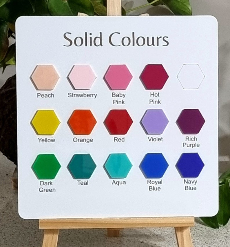 Op de afbeelding: Een wit bord met de tekst "Solid Colours" bovenaan. Het bord is verdeeld in drie rijen met zes zeshoekige kleurstalen. De kleuren zijn perzik, aardbei, babyroze, felroze, geel, oranje, rood, violet, donkerpaars, donkergroen, teal, aqua, koninklijkblauw en marineblauw.