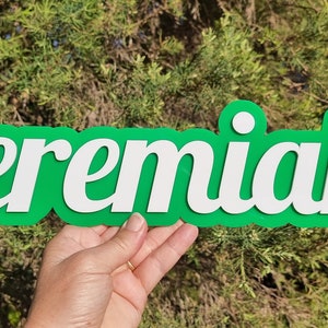 Op de afbeelding: Een groen en wit houten bord met de naam "Jeremiah" in witte letters. Het bord wordt in de hand van iemand gehouden.