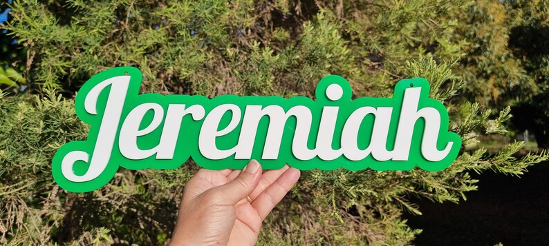 Op de afbeelding: Een groen en wit houten bord met de naam "Jeremiah" in witte letters. Het bord wordt in de hand van iemand gehouden.
