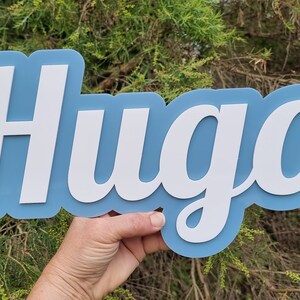 Op de afbeelding: Een blauw en wit houten naambord met de naam "Hugo" in een cursief schrift.