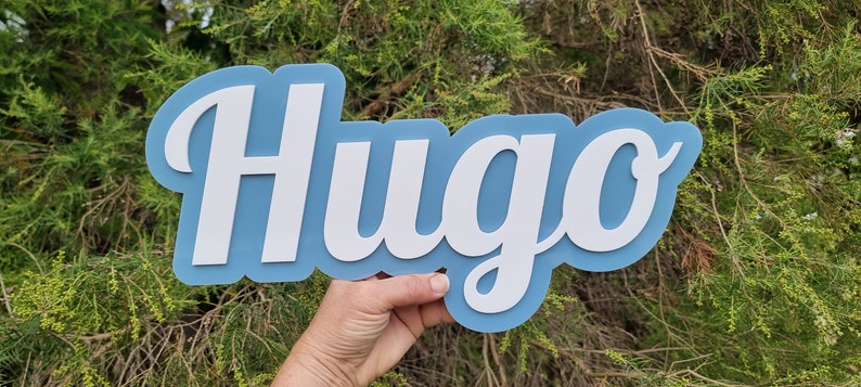 Op de afbeelding: Een blauw en wit houten naambord met de naam "Hugo" in een cursief schrift.