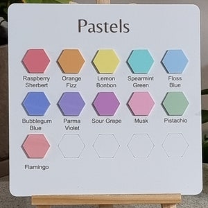 Pode incluir: Um quadro branco com a palavra "Pastels" no topo, exibindo uma paleta de cores de formas hexagonais em v&aacute;rios tons pastel. As cores incluem Raspberry Sherbert, Orange Fizz, Lemon Bonbon, etc. O quadro &eacute; apoiado em um pequeno cavalete de madeira.