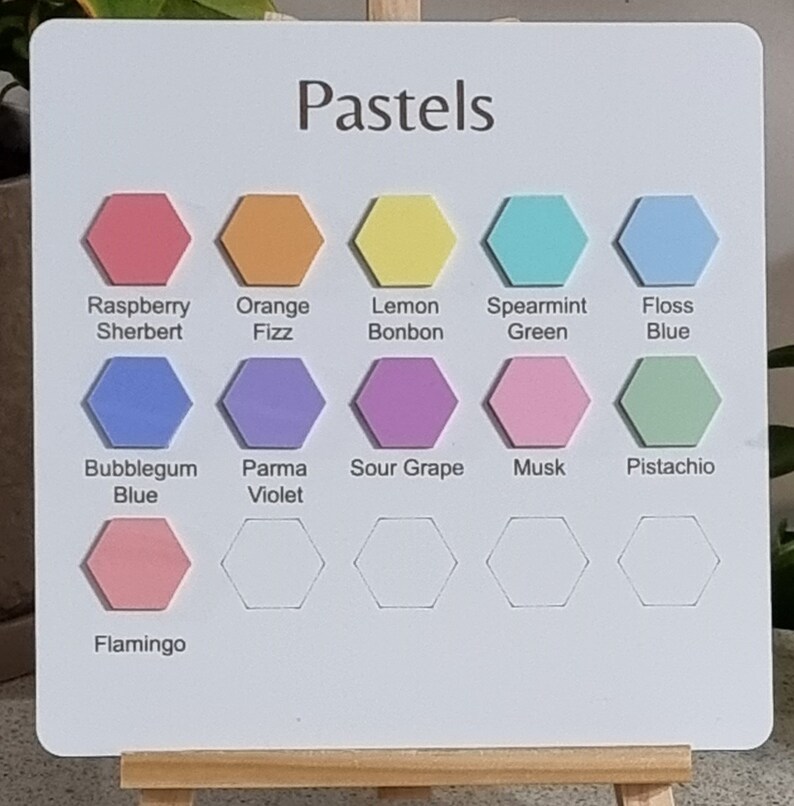Pode incluir: Um quadro branco com a palavra "Pastels" no topo, exibindo uma paleta de cores de formas hexagonais em v&aacute;rios tons pastel. As cores incluem Raspberry Sherbert, Orange Fizz, Lemon Bonbon, etc. O quadro &eacute; apoiado em um pequeno cavalete de madeira.