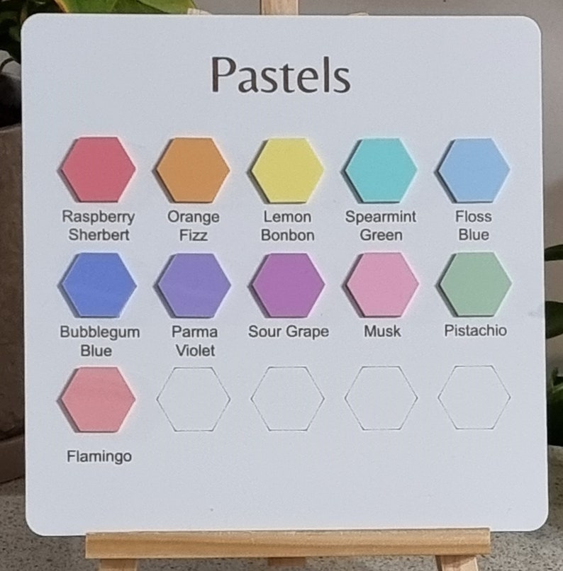 Op de afbeelding: Een wit bord met het woord "Pastels" bovenaan. Het bord is verdeeld in een raster met 12 zeshoekige vormen. Elke zeshoek is gevuld met een andere pastelkleur en gelabeld met een naam. De kleuren omvatten Raspberry Sherbert, Orange Fizz, Lemon Bonbon, Spearmint Green, Floss Blue, Bubblegum Blue, Parma Violet, Sour Grape, Musk, Pistachio en Flamingo.