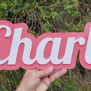 Op de afbeelding: Een roze en wit naambord met de naam "Charli" in witte cursieve letters.