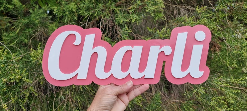 Op de afbeelding: Een roze en wit naambord met de naam "Charli" in witte cursieve letters.