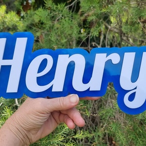 Op de afbeelding: Een blauw acryl bord met de naam "Henry" geschreven in witte cursieve letters.